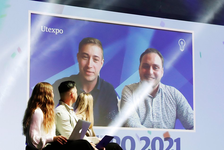 Bild på två personer på en stor skärm i bakgrunden. Till vänster sitter tre personer bortvända från kameran och tittar på personerna på skärmen. Foto.