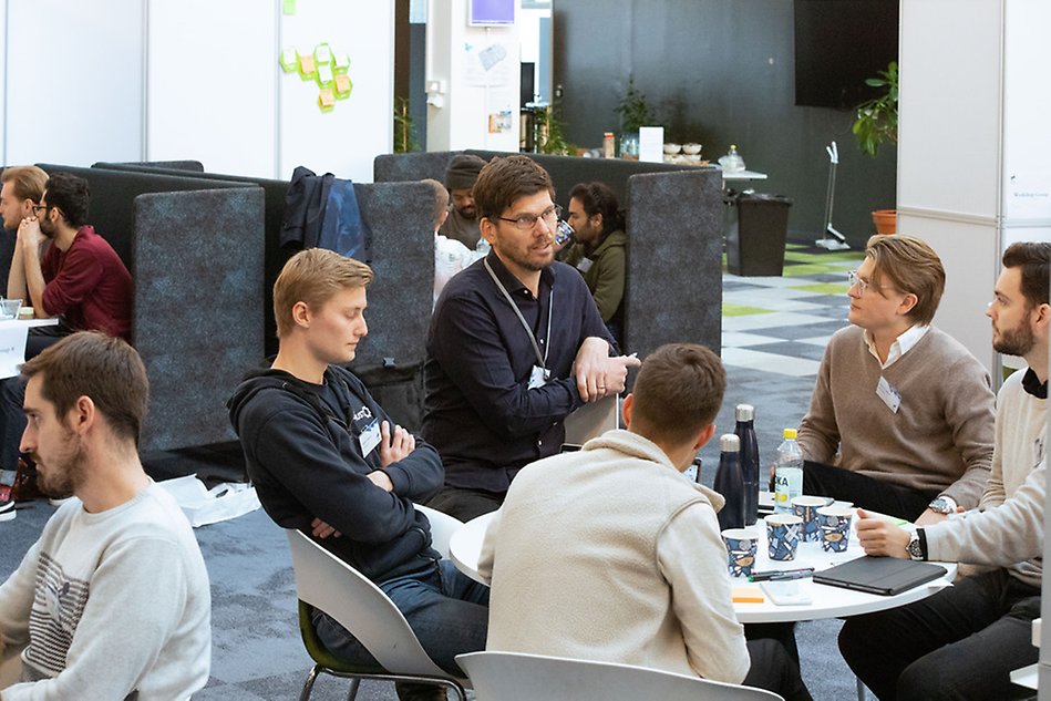 Four people sit around a table and talk to each other. Three of them are looking at the fourth person and one is looking down. Photo.