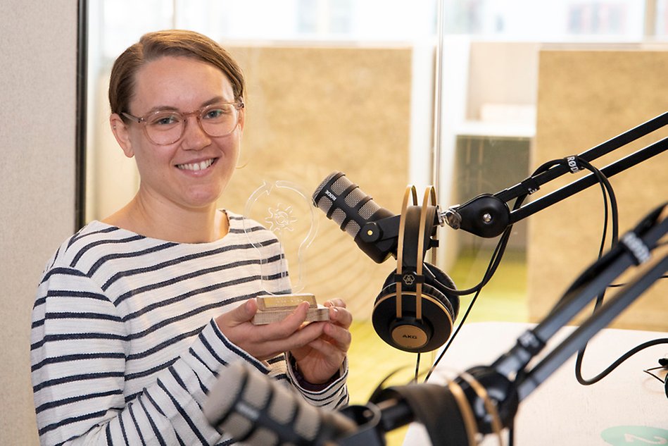 En kvinna i uppsatt hår och randig tröja står i en poddstudio bredvid en mikrofon. Foto.