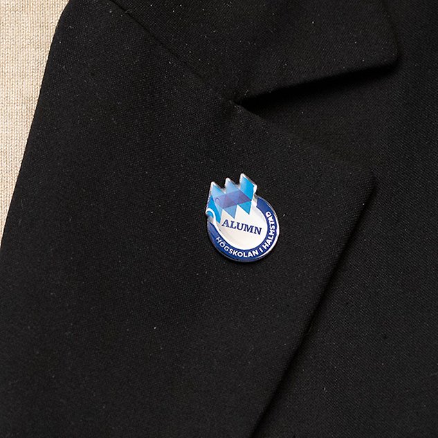 En pin med en logotyp och texten ”Alumn” syns på ett kavajslag. Foto. 