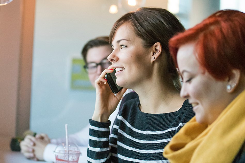 One man and two women sitting together by a table, smiling. One of them is talking on the phone. Photo.