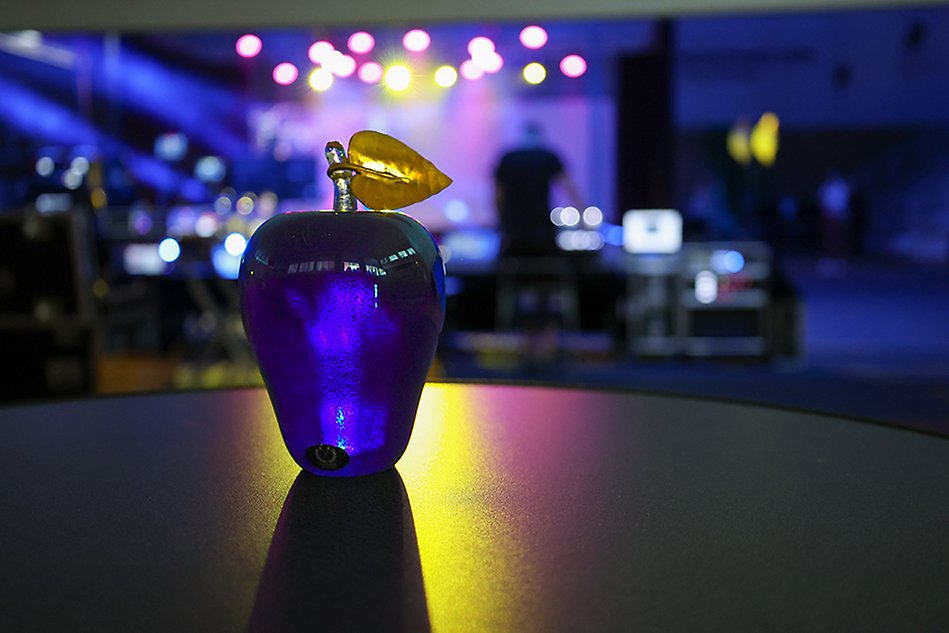 A blue apple-shaped glass ornament with a gold-colored leaf at the top sits on a table, a stage and spotlights are in the background. Photo.