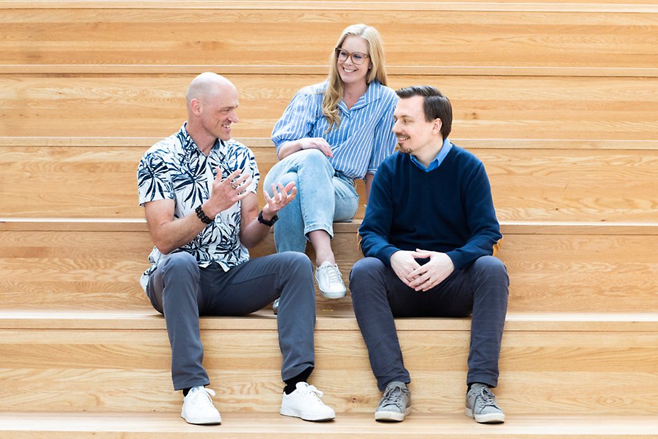 Three people in light-colored clothing are sitting on a staircase and conversing. Image.