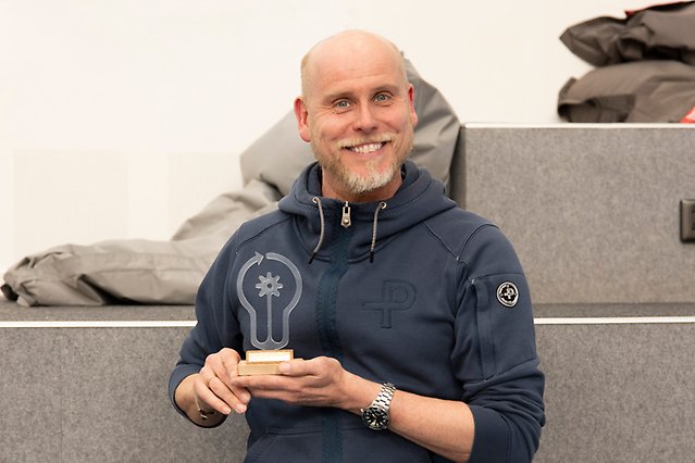 A man in a dark hoodie and white beard smiles to the camera and is holding an Utexpo statuette.