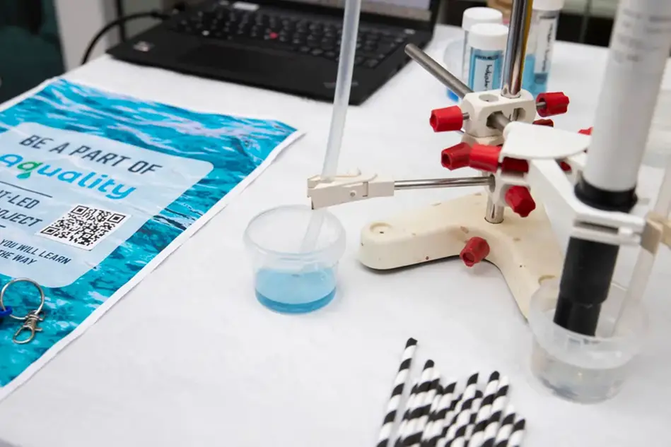 A device with a tube down into a plastic jar with colored liquid. Next to a poster that says aquality on it.