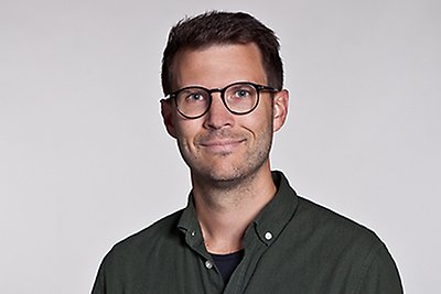 En person med mörkgrön skjorta, mörkt hår och runda glasögon ler mot kameran. Porträttbild. 