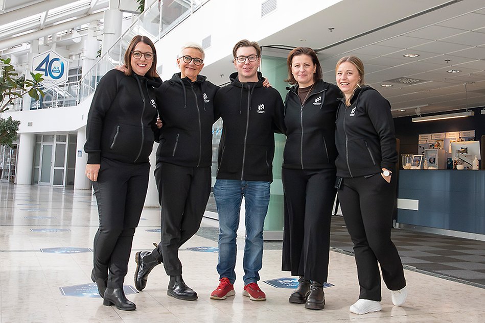 Five people are standing in a row with their arms around each others shoulders in a bright environment indoors. They all have black shirts and are looking at the camera smiling. Photo. 