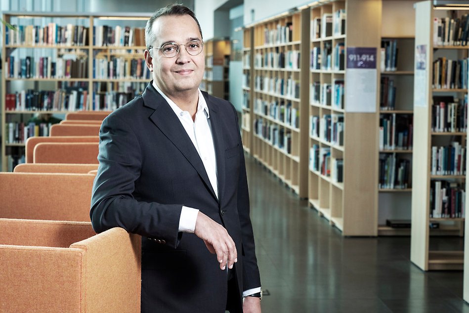 A man wearing a dark jacket standing, leaning with his arm against a char in a room full of books. Photo.