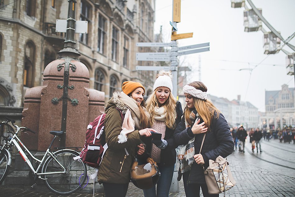 Three young women. in winter clothes and with bags, in front. Two women looking down in a cell phone, one looking up. Building and people in the background. Likely the women are tourists in a big city. Phot.