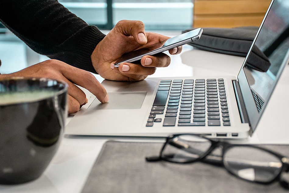 Close up photo of a person’s hands using phone and laptop. In the front a cup with beverage of some kind and a pair of glasses.