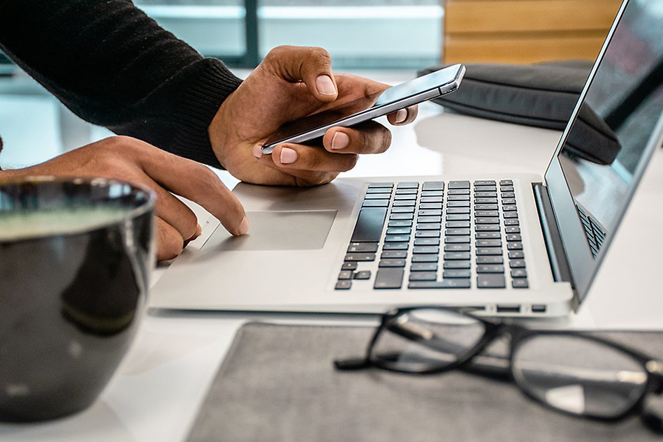 Close up photo of a person’s hands using phone and laptop. In the front a cup with beverage of some kind and a pair of glasses. 