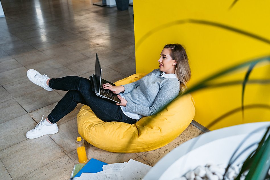 A young woman sitting on a big yellow pillow on the floor with a laptop on her knee. She is looking at it and typing. Phot from the side