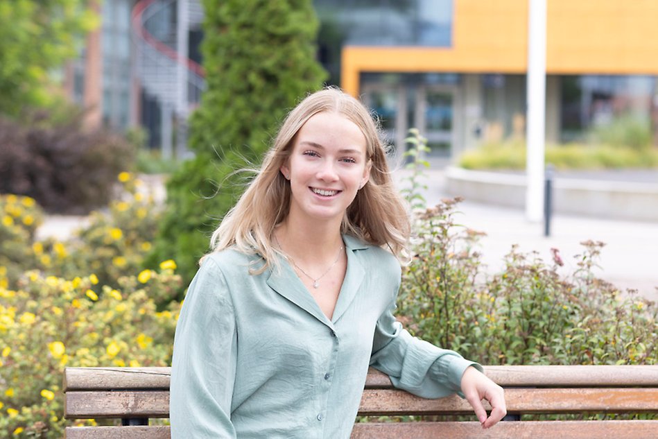 Blond tjej i pastellgrön blus sitter ner på en bänk och ler mot kameran. I bakgrunden finns entrén till hus S. Foto.