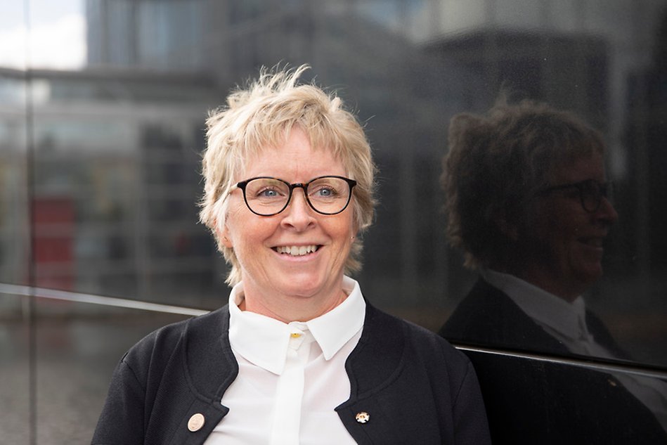 A person with short hair and glasses smiles. The person looks beyond the camera, and there is a black background. Photo.