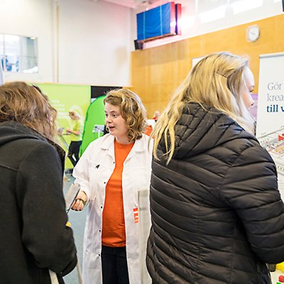 En person i vit rock och orange tröja tittar på och ser ut att prata med en person till vänster. Foto.
