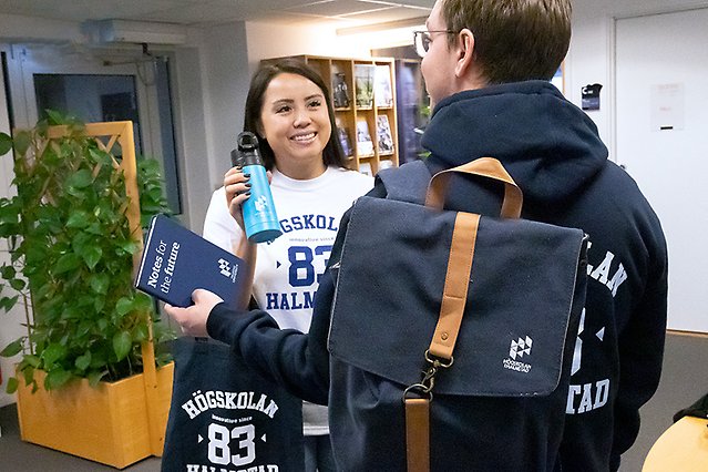 Två personer där man ser den ena framifrån tittar på varandra och ler. De bär kläder och andra profilprodukter där det står ”Högskolan 83 Halmstad”. Foto.