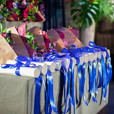Rolled-up papers with blue, shiny ribbons are lined up on a table along with bouquets of flowers. Photo.