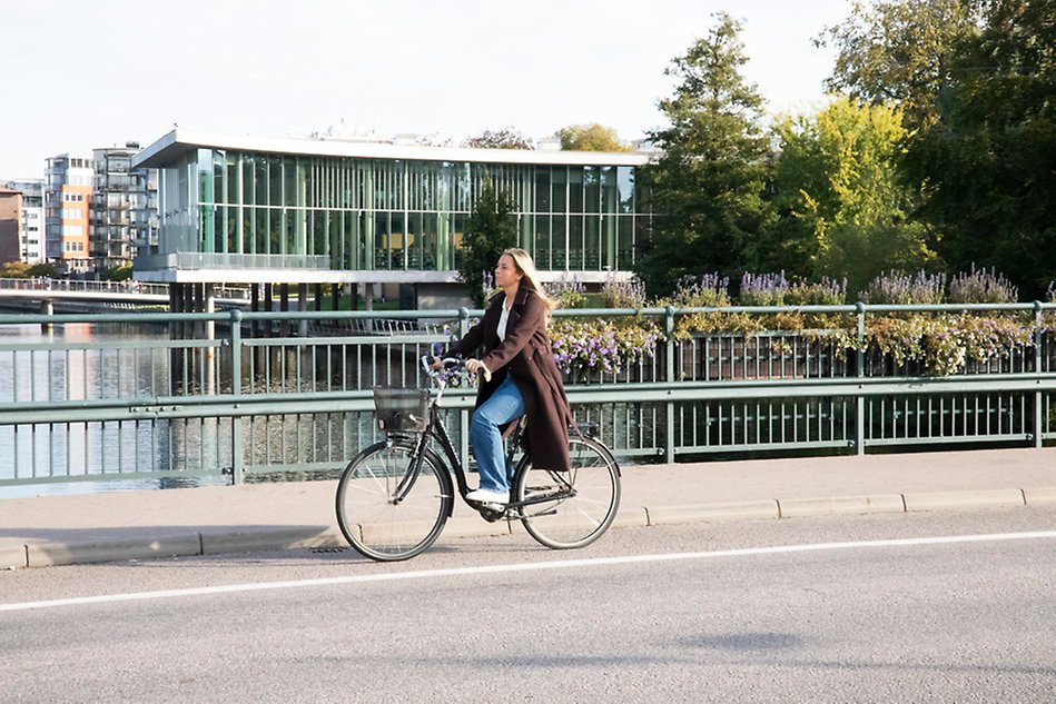 A young woman biking on a bridge with a modern building and a river in the background. Photo.