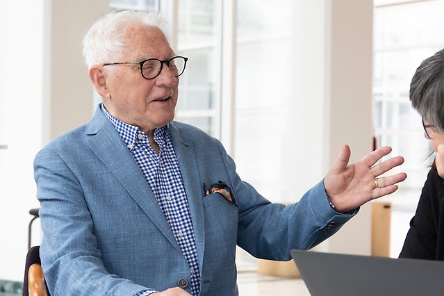 An elderly man in a blue jacket is seen on the left in profile talking to a person on the right of whom you can only see a glimpse. Photo.