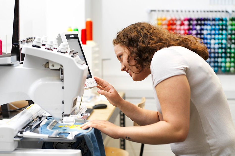 En person med långt, lockigt hår och vit t-shirt står framåtböjd över en symaskin.