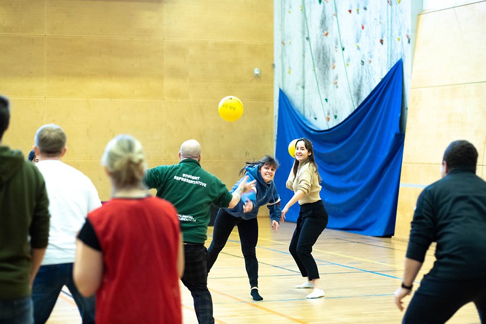En grupp unga människor i en idrottshall leker en lek där de kastar mjuka gula bollar på varandra. I fokus är två kvinnor som kastar bollar. Foto.