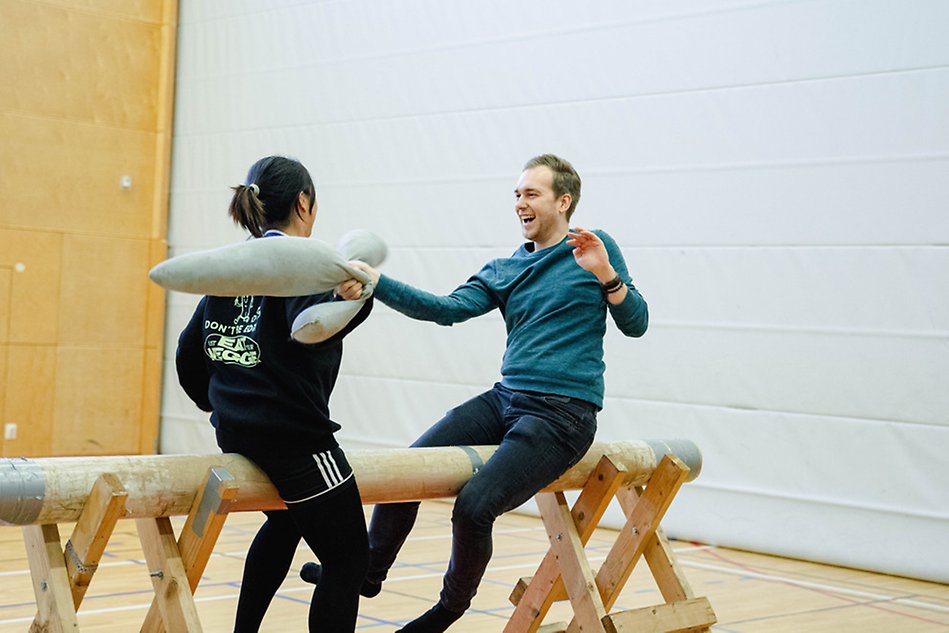 En kvinna och en man sitter på en gymnastikbock i en idrottshall och slår mot varandra med mjuka kuddar. Mannen till höger i bilden har ett stort leende. Foto.