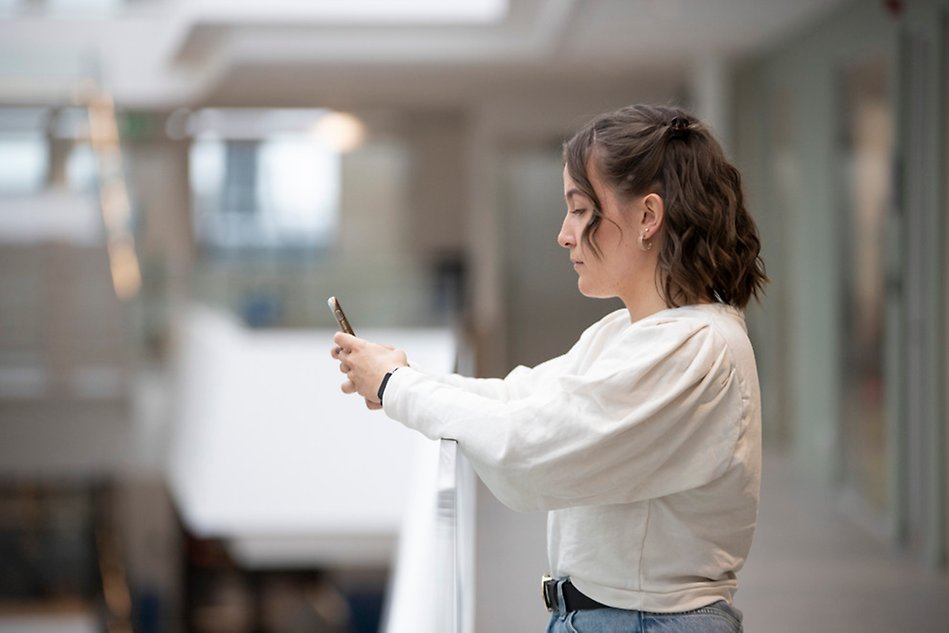 A person wearing a white shirt leaning against a handrail, holding and looking at a smartphone.