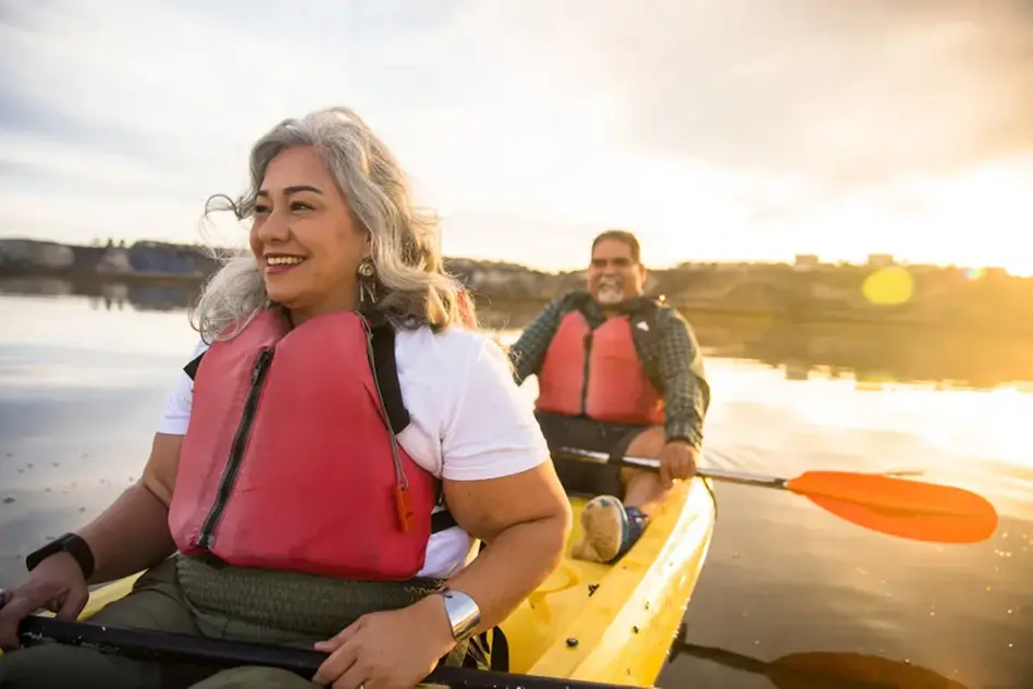 Two people in a kayak. They are smiling and the sun is setting across the water. Photo.