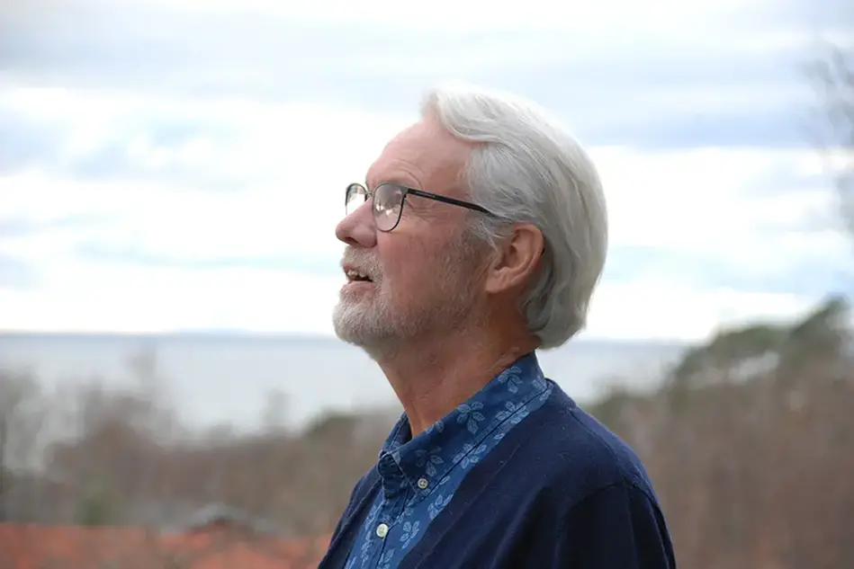 A man with white hair and black glasses standing outside looking up, in profile. Photo.