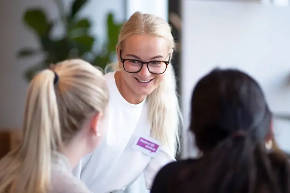 En ung man med skägg sitter vid ett bord intill ett fönster. Han ser in i kameran och ler. Foto.