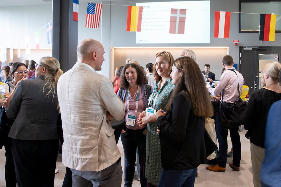 A group of people stand and chat beneath a ceiling decorated with flags from various countries. Photo