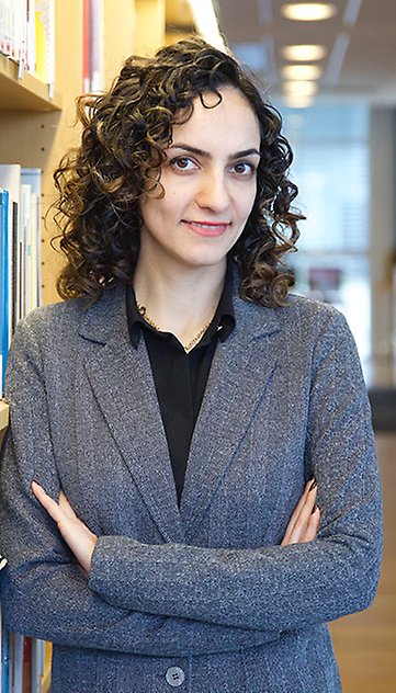 A smiling woman with curly dark hair and folded arms leaning against a bookcase. Photo.