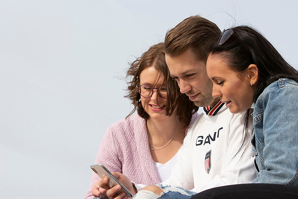 Tre unga personer sitter bredvid varandra med gråblå himmel i bakgrunden och tittar ner i en mobiltelefonskärm. Foto.