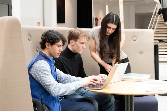 Three students studying in front of a laptop in a campus environment. Photo.