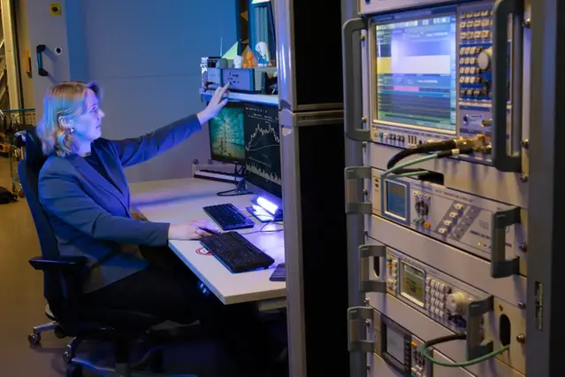 A woman is sitting at a desk in an electronics lab, with a computer screen in front of her. One hand is placed on a mouse and the other one is pressing a button on a machine above the screen. Photo.