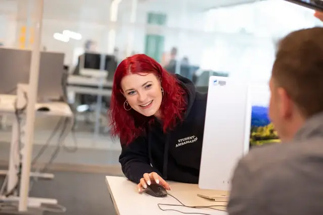 A student peeks out from behind a screen while her hand rests on a computer mouse. Photo.