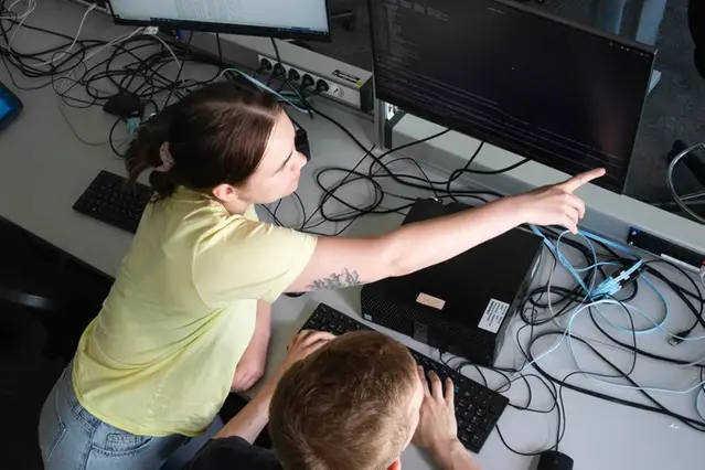 One person seen from above. pointing at a computer screen, while bending over a table full of cables. Photo.