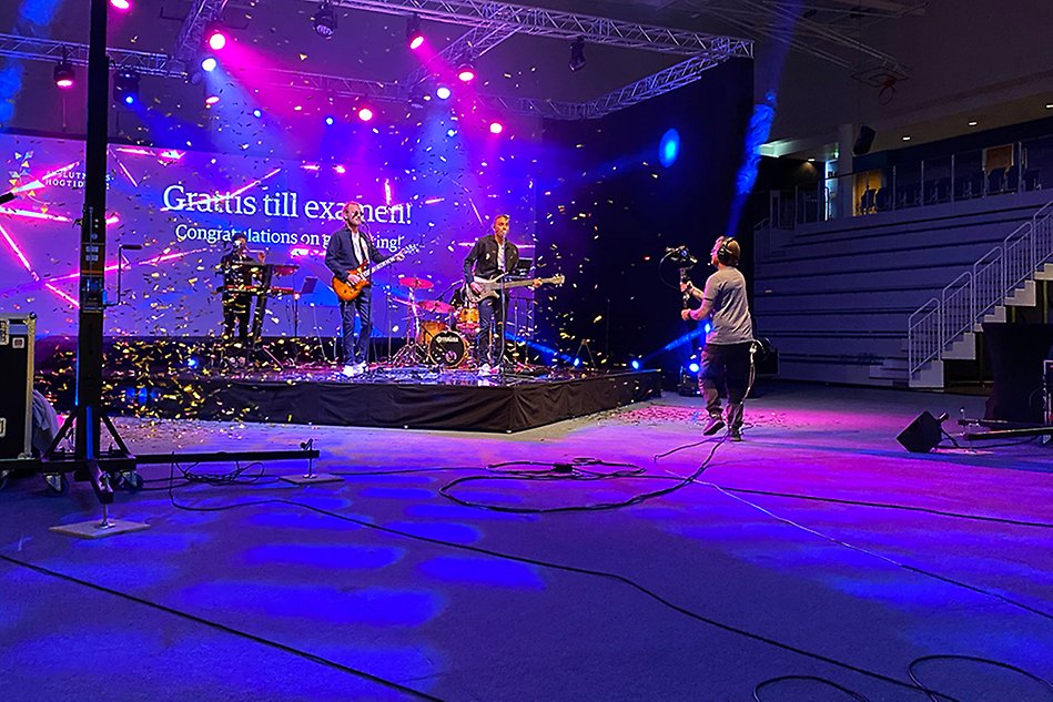 A music group performing on a stage in front of a large screen while it rains confetti across the stage. A cameraman can be seen in the foreground. Photo.