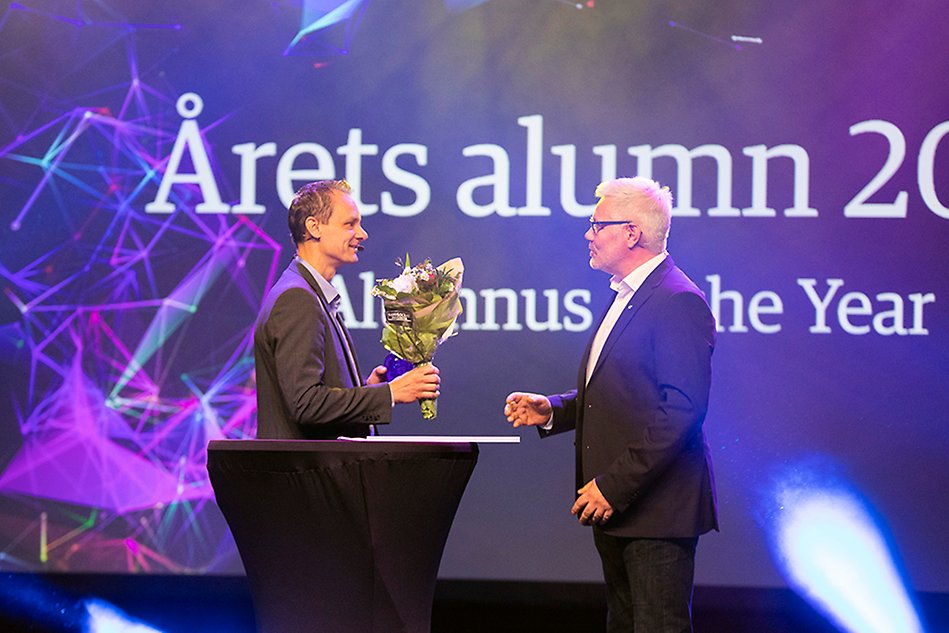 Two people facing each other in front of a large, colourful screen. The person on the left is holding a bouquet of flowers. Photo. 