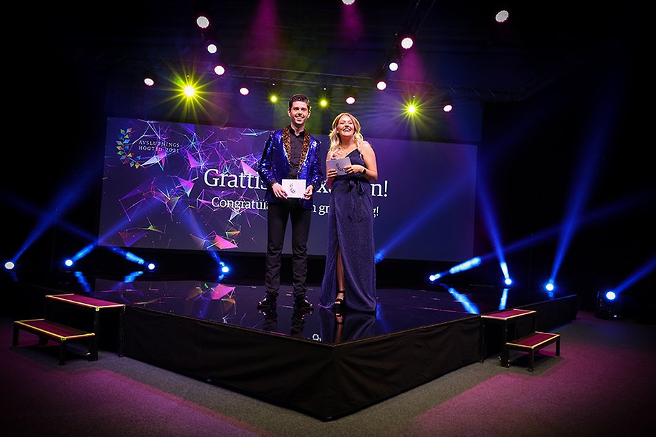 A man and a woman dressed in festive clothes stand on a stage lit up by spotlights. Photo.