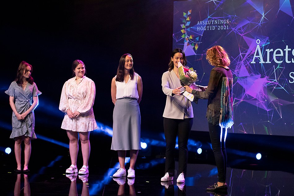 Four young women stand in a row on a stage, one of them is receiving flowers from another woman. Photo.