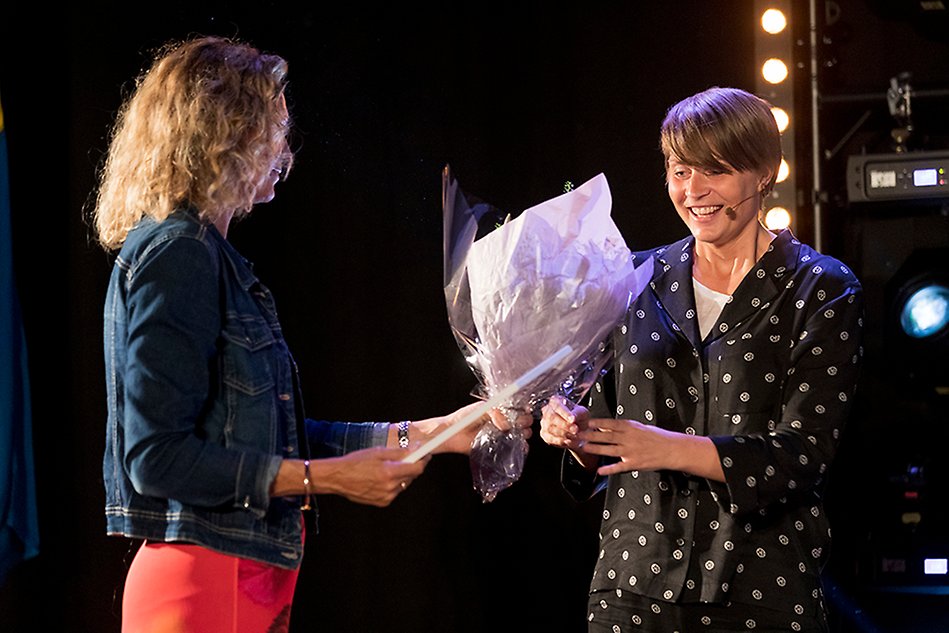 Two women standing in a spotlight, one of them handing flowers and a diploma to the other one. Photo. 