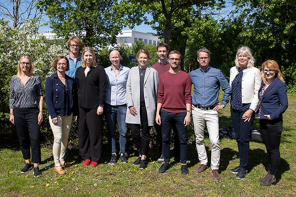 De elva projektdeltagarna står i solen på en grön gräsmatta framför blommande buskar. Foto.