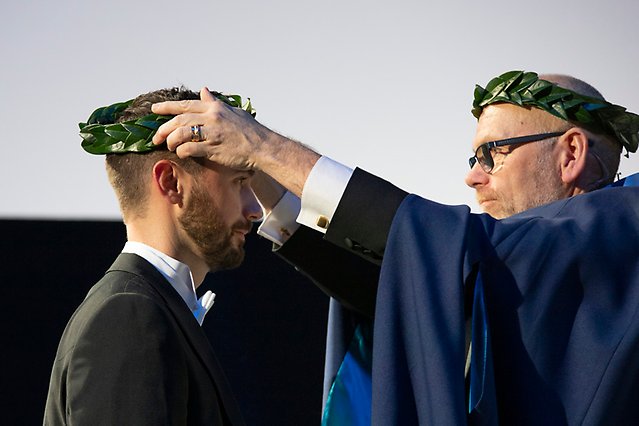 A man with a wreath on his head, dressed in a cape seen in profile, puts a wreath on another man’s head. Photo
