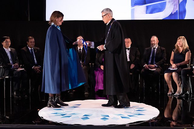 A man hands over a diploma to a woman. In the background, a group of people sit in a semicircle. Photo
