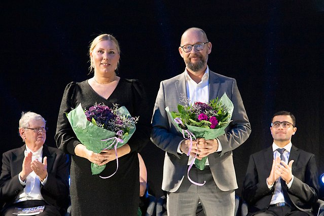 A woman and a man stand with a bouquet of flowers in their hands and look into the camera. Behind them are two men applauding. Photo