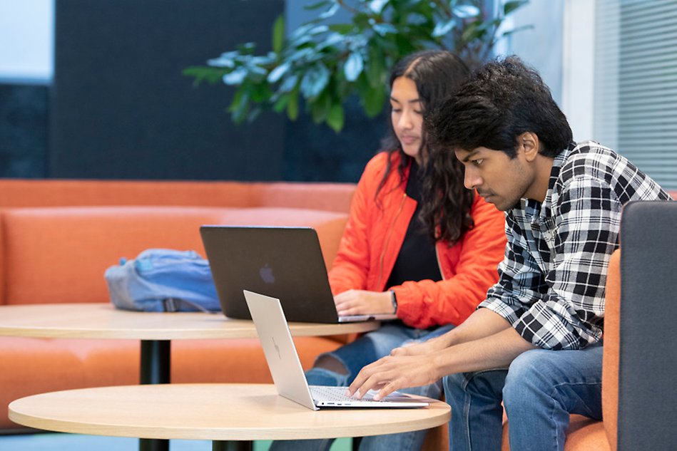 A young woman and a young man are sitting next to each other on a sofa. They each have a laptop in front of them on a small round table. Photo.
