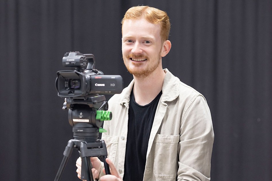 Rödhårig kille i beige, öppen skjorta och svart t-shirt håller i en filmkamera och ler mot kameran. Foto.