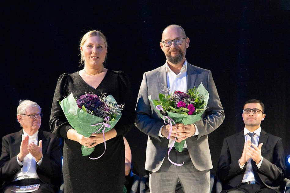 En kvinna och en man står med blombukett i händerna och tittar in i kameran. Bakom dem syns två män som applåderar. Foto