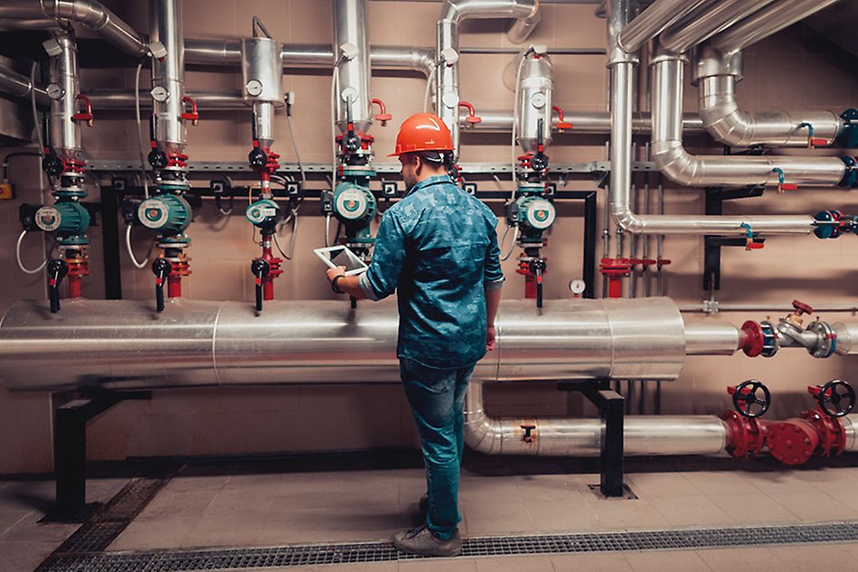 A man wearing a safety helmet and holding a tablet standing in front of a wall with pipes and valves. Photo.